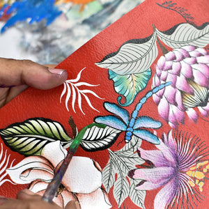 A person's hand painting a floral design on a textured red surface of an Anuschka Medium Zip-Around Eyeglass/Cosmetic Pouch - 1163.