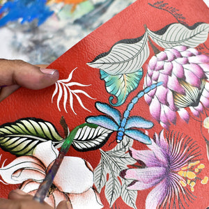 A person's hand painting a colorful floral design on a red textured Anuschka Card Holder with Wristlet - 1180 with RFID protection.