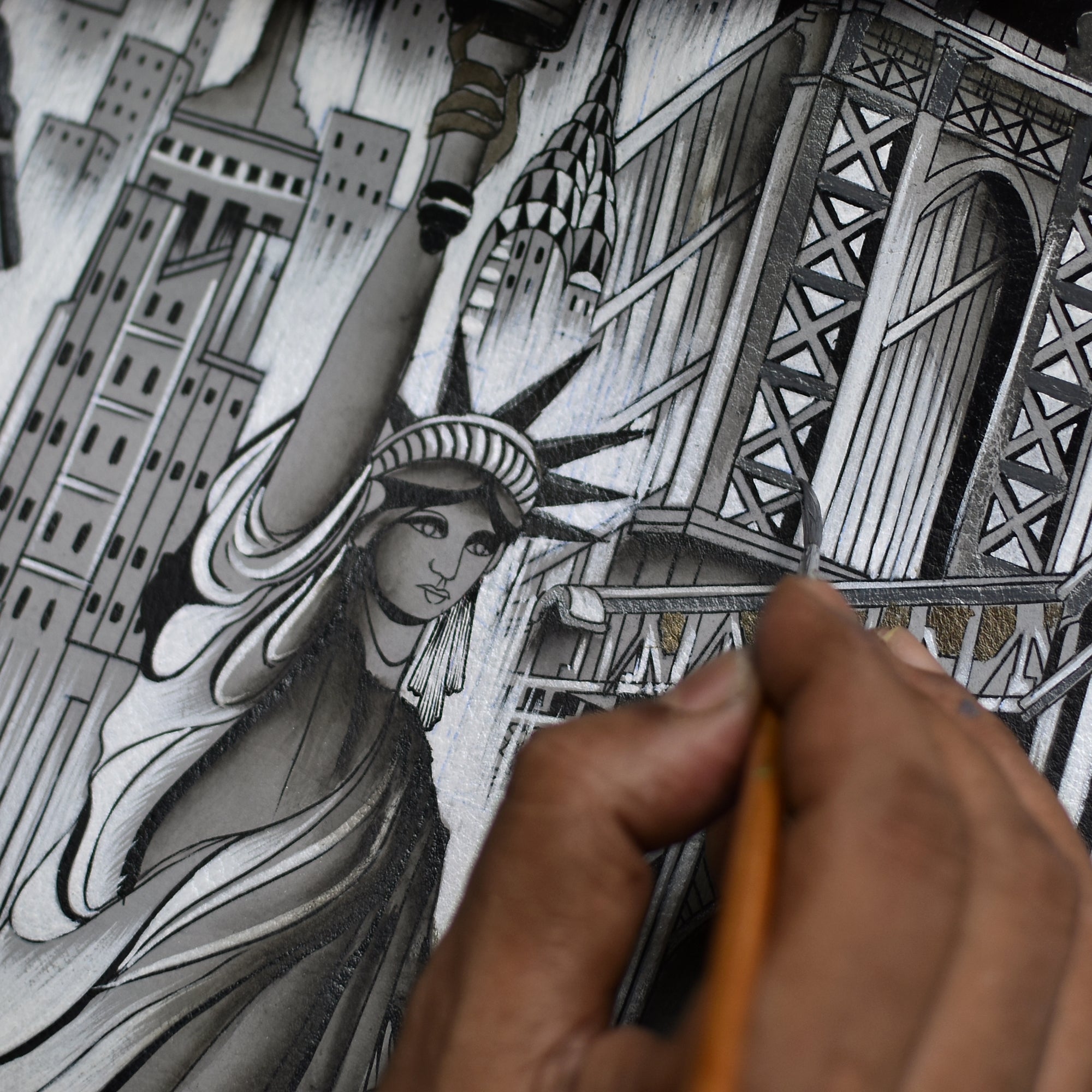 Close-up of a hand painting a detailed black and white illustration of the Statue of Liberty and surrounding New York City skyscrapers, with the Anuschka Slim Shoulder Hobo - 714 lying open nearby.