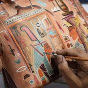 A person meticulously painting ancient Egyptian figures and hieroglyphics on a spacious Anuschka Expandable Shopper Tote - 712, using a small brush.