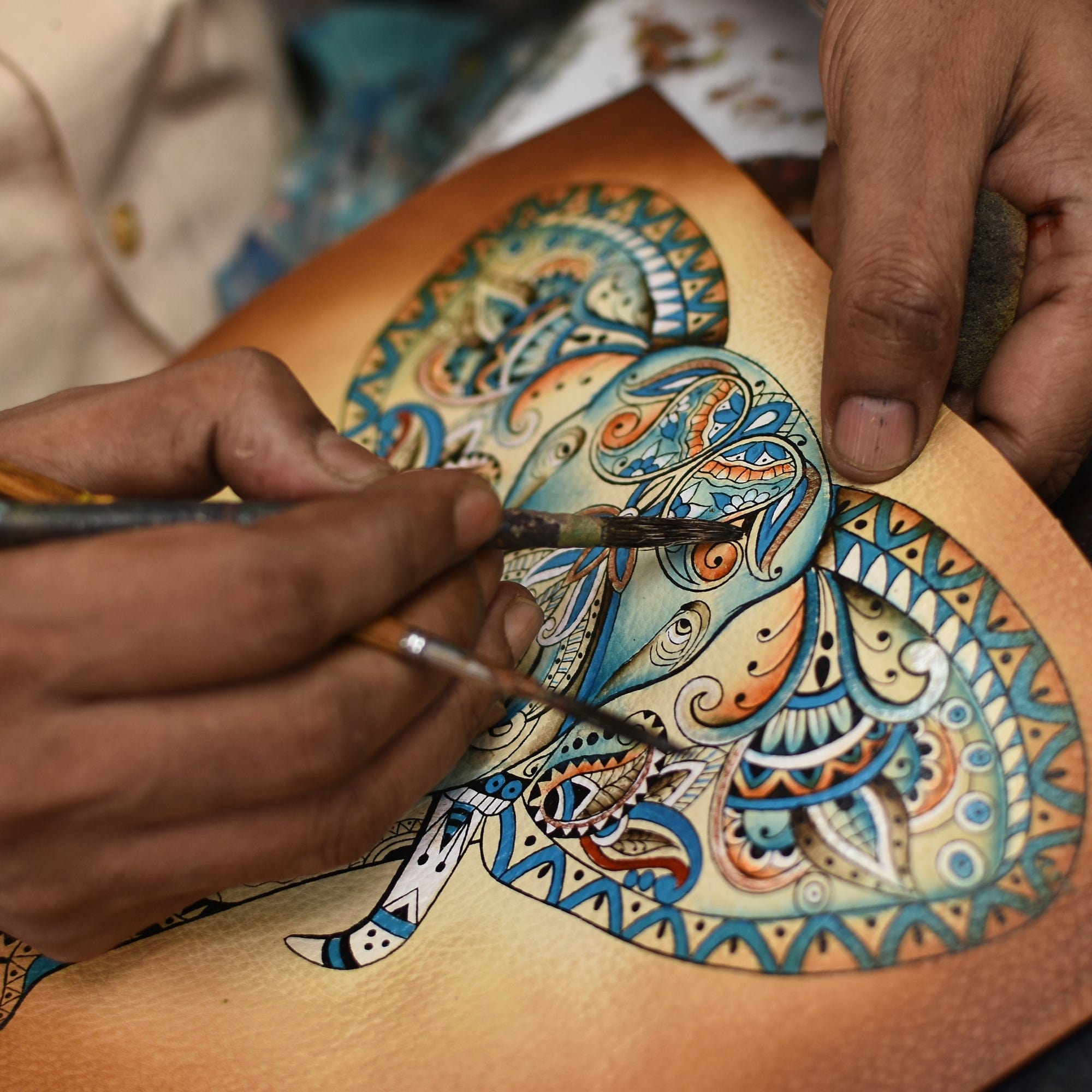 A person uses paintbrushes to add intricate designs and colors to a detailed, hand-painted, decorative painting of an elephant on the Anuschka Classic Hobo With Side Pockets - 382.