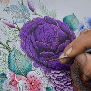 Artist's hand using a fine brush to add details to a purple flower illustration on an Anuschka Crossbody With Front Zip Organizer - 651 with an adjustable strap.