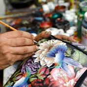 Close-up of an artist's hand painting a detailed floral and bird design on genuine leather with a fine brush, surrounded by jars of paint, Crossbody Phone Case - 1173 by Anuschka, crossbody straps, and RFID card holders in the background.