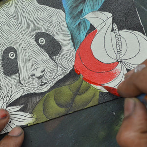 An artist's hand painting a red and green butterfly on a canvas featuring a Anuschka Medium Zip Pouch - 1107 with a black and white panda illustration.
