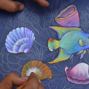 Hands coloring a chic marine life sketch with vibrant hues on a blue canvas using the Anuschka Medium Satchel - 697.
