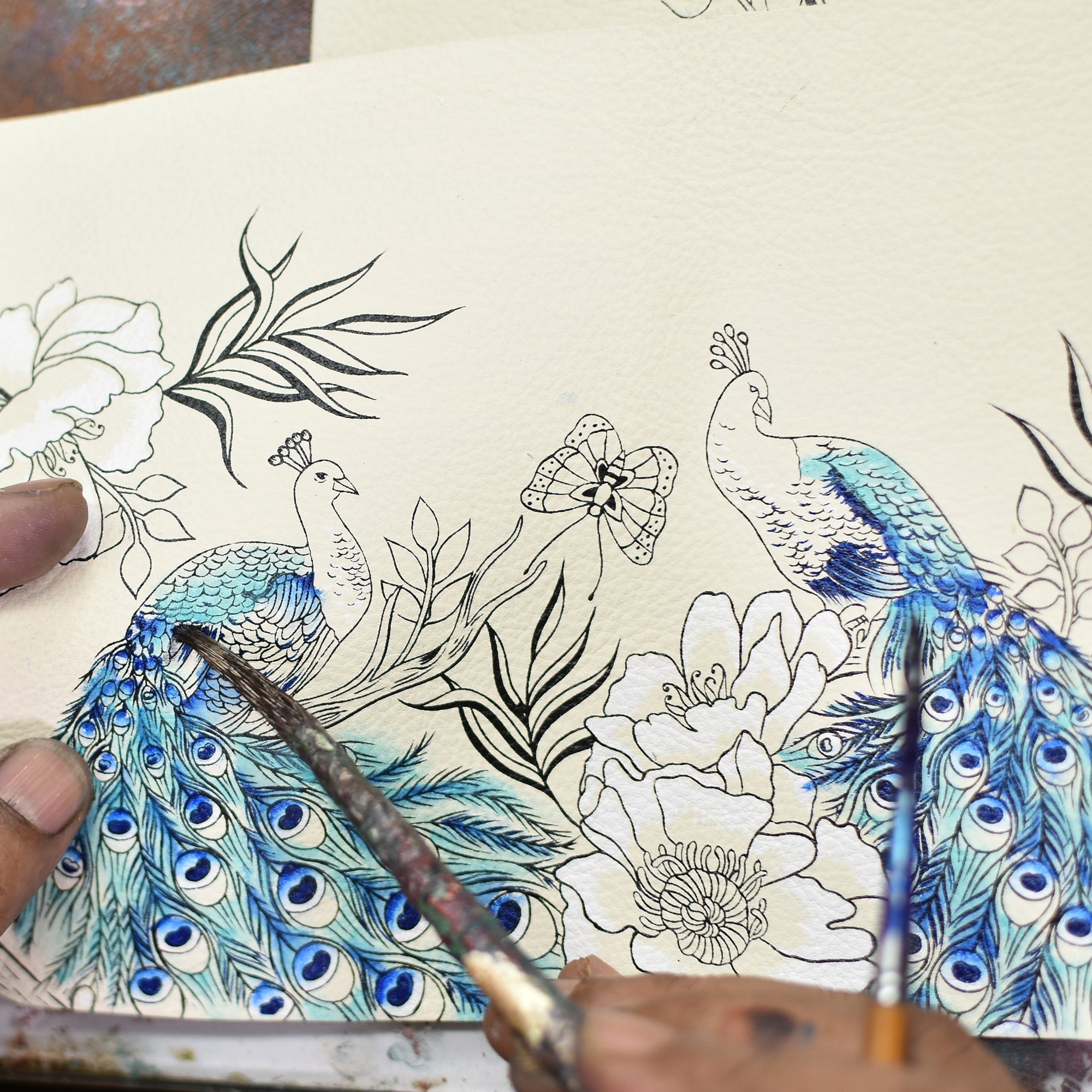 Hands painting intricate peacock illustrations on a genuine leather surface of the Anuschka Airpod Pro Case - 1179, adding blue hues to the feathers and detailing flowers and leaves around the birds.