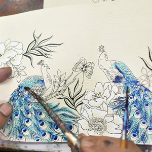 An artist's hands holding a paintbrush, adding blue details to an intricately designed peacock illustration on an Anuschka Accordion Flap Wallet - 1112.
