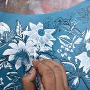 A person is painting a detailed floral and butterfly design in white on a blue surface using a fine brush, reminiscent of the intricate patterns found on an Anuschka Accordion Flap Wallet - 1174 made from genuine leather. The background has a textured, multicolored surface.