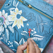 Close-up of a person's hand painting flowers and leaves on a blue genuine leather Clasp Pouch With Key Fobs - 1177 by Anuschka using a small brush. The pouch, with its vintage charm, features a dragonfly and floral design in various stages of completion.