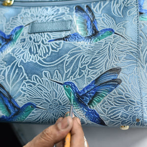 Hand painting a hummingbird design on a blue Anuschka Medium Zip Pouch - 1107 genuine leather purse.