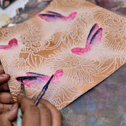 An artist's hand applying pink and purple paint to a textured canvas with floral designs on an Anuschka Medium Crossbody With Double Zip Pockets - 447 genuine leather travel easel featuring an adjustable shoulder strap.