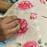 A person is painting embossed roses and butterflies on a decorative surface of the Two Fold Organizer Wallet - 1178. The roses are varying shades of pink and red, beautifully contrasting with the light cream color of the genuine leather, perfect for minimalists. The wallet is a product from Anuschka.