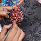 Close-up of hands painting a delicate red rose on the black embossed surface of the Anuschka Two Fold French Wallet - 1181, which features intricate roses and butterflies. A paintbrush meticulously applies color to the detailed floral design, transforming this hand-painted leather wallet.