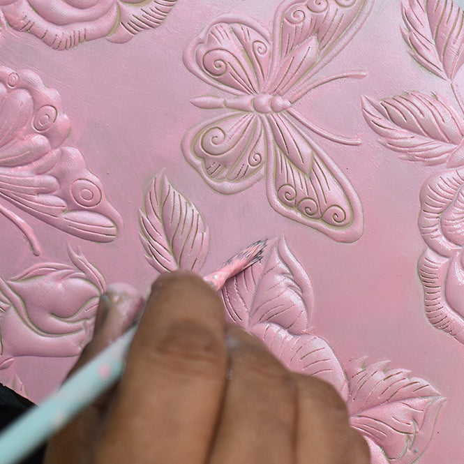 A person applies pink paint on a textured surface featuring butterfly and floral designs, creating an exquisite custom look perfect for embellishing the Anuschka Crossbody Phone Case - 1173.