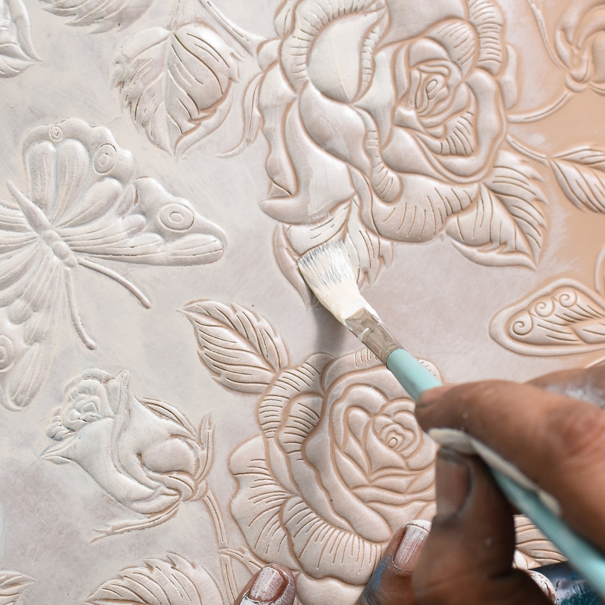 A minimalist artist meticulously paints details on a Two Fold Organizer Wallet - 1178 featuring raised floral and butterfly designs using a paintbrush with white paint by Anuschka.