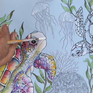 A hand using a paintbrush to color an illustrated sea turtle, with jellyfish and coral in the background, creating stunning hand-painted artwork on an Anuschka RFID Blocking Small Flap French Wallet - 1138.