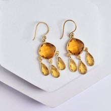 Load image into Gallery viewer, Gold-tone Vanya Lara Triple Dew Drop Earrings with amber-colored hydro stone setting on a white display stand.
