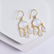 A pair of 22k gold-plated earrings with large, faceted white Hydro Quartz gemstones and three smaller teardrop-shaped gemstones hanging from each. The Triple Dew Drop Earrings - VER0006 by Vanya Lara are displayed on a light grey geometric surface.