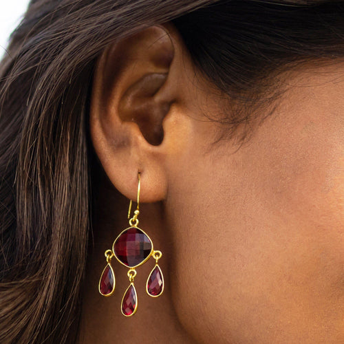 A close-up of a woman's ear wearing Vanya Lara's Triple Dew Drop Earrings with red gemstones in a gold setting.