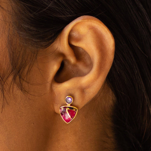 A close-up of a person's ear showcasing a triangle drop earring with gold plating and a small gemstone at the top by Vanya Lara.