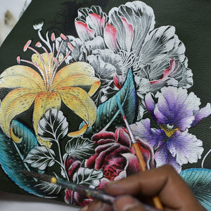 An artist's hand painting intricate floral designs on an Anuschka Medium Zip Pouch - 1107.
