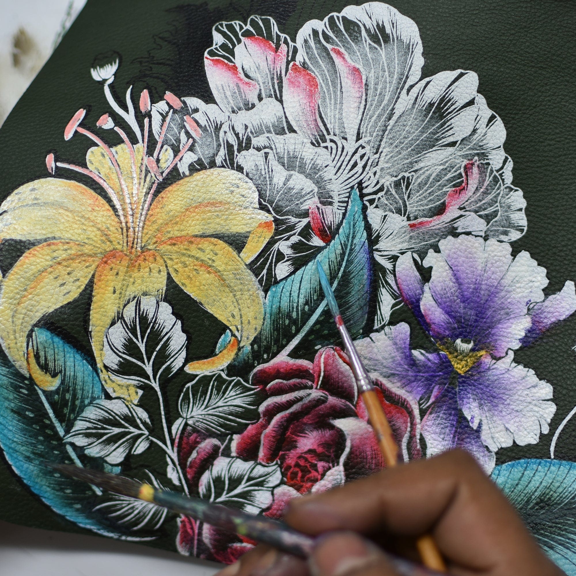 An artist's hand meticulously paints vibrant flowers, including a yellow lily and purple blooms, on the dark genuine leather surface of the exquisite Anuschka Zip-Top Shoulder Hobo - 707. Brushes and detailed floral patterns adorn the front of this elegant hobo bag, which features a chic chain detail shoulder strap.