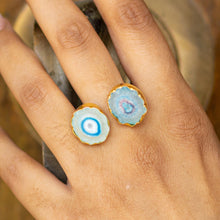 Load image into Gallery viewer, A hand displaying two Solar Quartz Rings from the Vanya Lara ring collection with blue agate gemstones against a natural backdrop, now available with free shipping.
