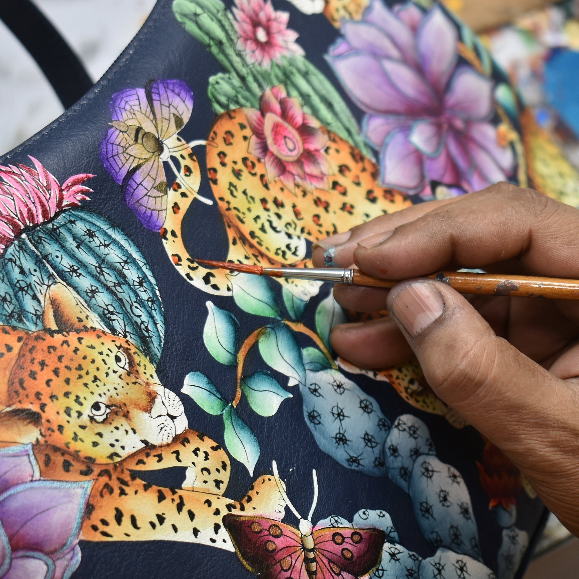 A person painting a colorful illustration on a dark surface featuring leopards, flowers, cacti, and butterflies using a fine brush, with their Anuschka Expandable Shopper Tote - 712 nearby to hold all their artistic supplies.