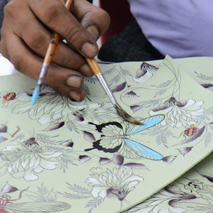 An artist's hands painting a butterfly on a floral-patterned Anuschka Wide Organizer Satchel - 695.