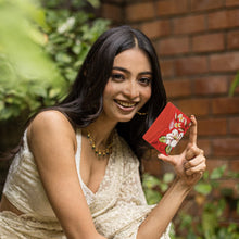Load image into Gallery viewer, Woman smiling outdoors and showing a small leather credit card case with hand-painted floral artwork by Anuschka.
