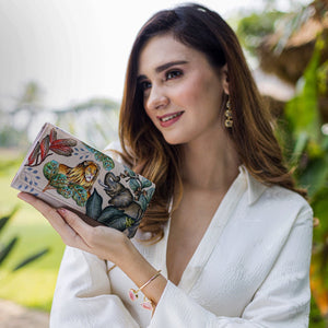 A woman in a white outfit holding an Anuschka Accordion Flap Wallet - 1112 with RFID protection, featuring a floral and animal design.