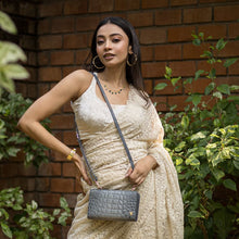 Load image into Gallery viewer, A woman in a cream dress posing with an Anuschka Organizer Wallet Crossbody - 1149 against a brick wall with green foliage.

