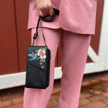 Load image into Gallery viewer, A person in a pink suit holding a black floral embroidered genuine leather Anuschka Crossbody Phone Case - 1173.
