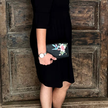 Load image into Gallery viewer, A person wearing a black dress with a floral pattern on the pocket and holding an Anuschka Accordion Flap Wallet - 1174 with RFID protection, standing against a wooden door.
