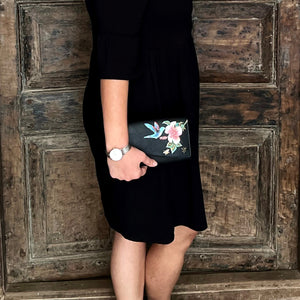 A person wearing a black dress with a floral pattern on the pocket and holding an Anuschka Accordion Flap Wallet - 1174 with RFID protection, standing against a wooden door.