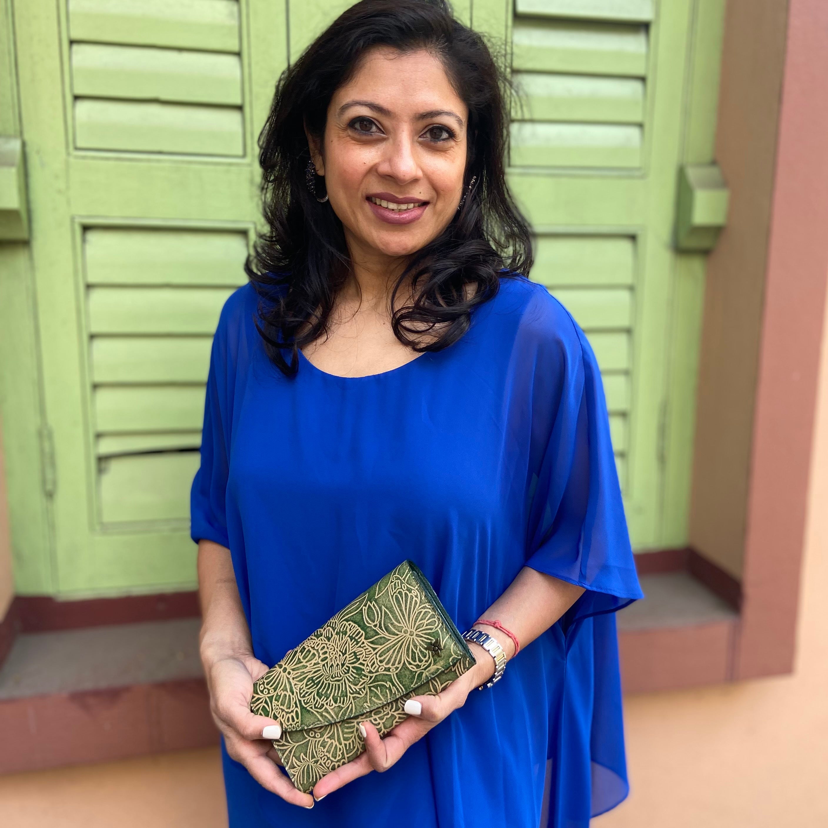 Woman in a blue dress holding an Anuschka Accordion Flap Wallet - 1174 with RFID protection, standing in front of green doors.