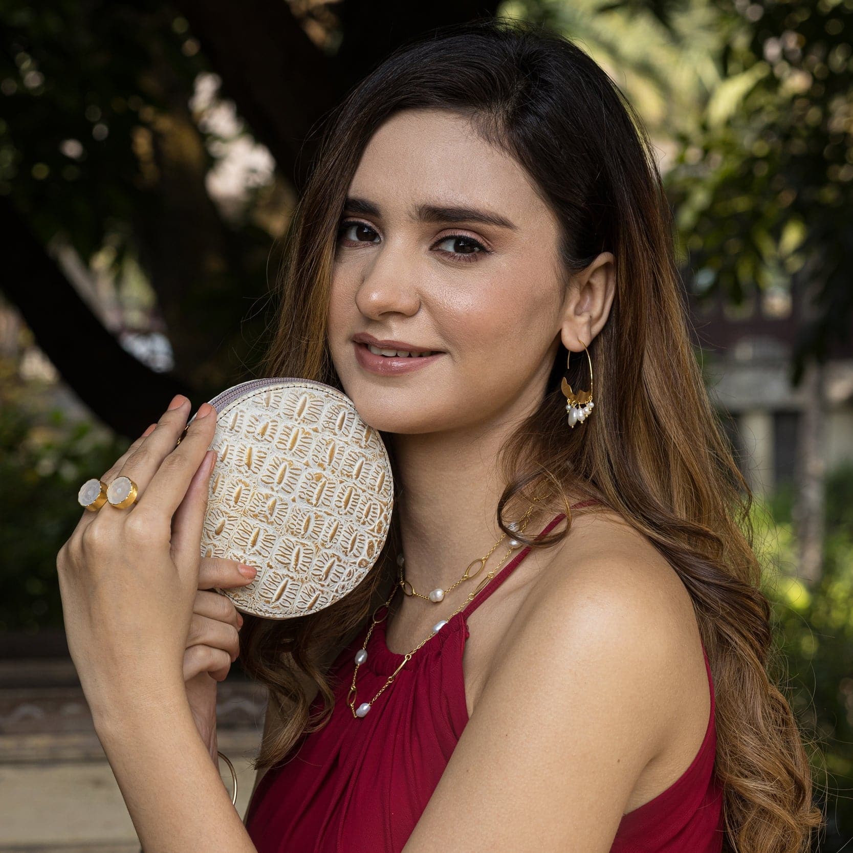 A woman with makeup, holding an Anuschka Round Coin Purse - 1175, and wearing a red dress accessorized with earrings and rings.