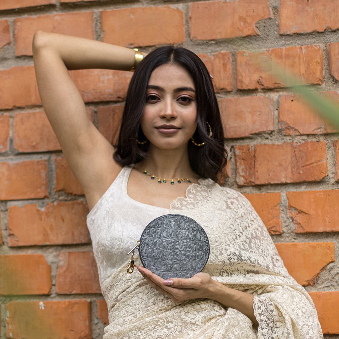 A woman in a lace dress posing with one hand behind her head and holding an Anuschka Round Coin Purse - 1175 against a brick wall background.