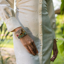 Load image into Gallery viewer, Person wearing a traditional cream-colored outfit with a detailed, Anuschka Adjustable Leather Wrist Band - 1176 made of genuine leather, standing in a garden setting.
