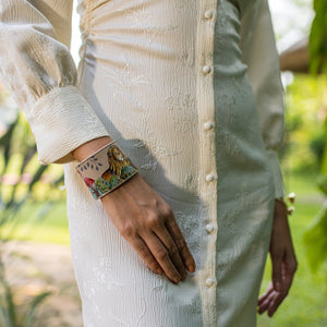 Person wearing a traditional cream-colored outfit with a detailed, Anuschka Adjustable Leather Wrist Band - 1176 made of genuine leather, standing in a garden setting.