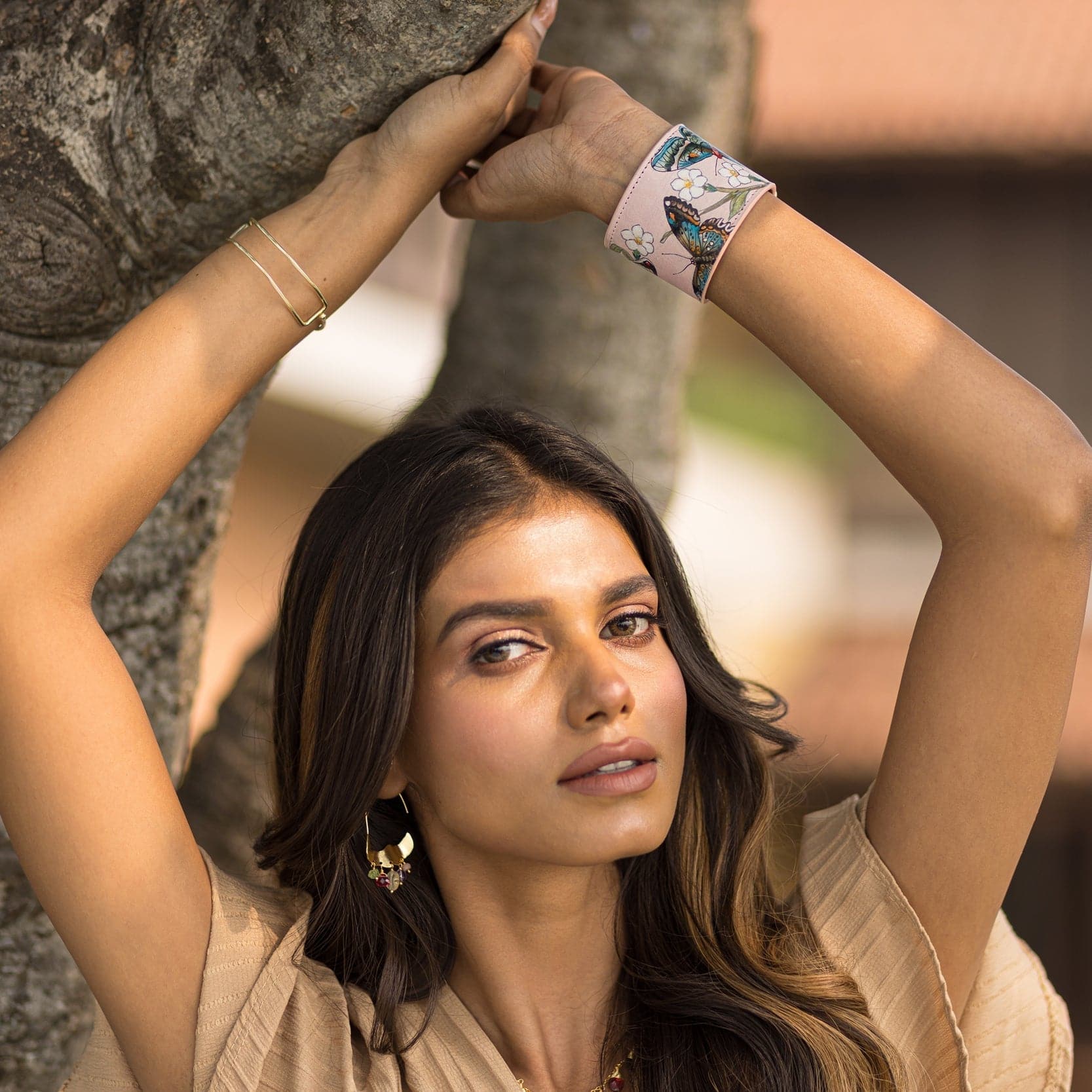 A woman with long hair, wearing earrings and an Anuschka Leather Adjustable Leather Wrist Band - 1176, leans against a tree.