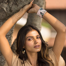 Load image into Gallery viewer, A woman with long hair, wearing earrings and an Anuschka Leather Adjustable Leather Wrist Band - 1176, leans against a tree.
