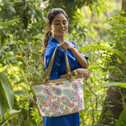 Woman in a blue dress holding an Anuschka Fabric with Leather Trim Zip Top City Tote - 12005 handbag with a leather shoulder strap in a lush garden setting.