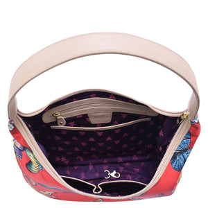 An open, patterned Anuschka handbag with a cream-colored shoulder strap and a purple interior displaying zippered pockets and brand logo.