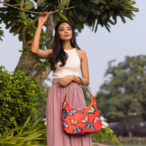 A woman holding a branch and carrying an Anuschka Fabric with Leather Trim Large Sling Hobo - 12010 bag with zippered pockets stands outdoors.