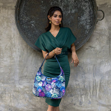 Load image into Gallery viewer, A woman in a green dress stands against a textured wall holding an Anuschka Fabric with Leather Trim Large Sling Hobo - 12010 with a zippered pocket.

