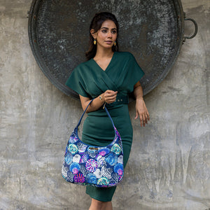 A woman in a green dress stands against a textured wall holding an Anuschka Fabric with Leather Trim Large Sling Hobo - 12010 with a zippered pocket.