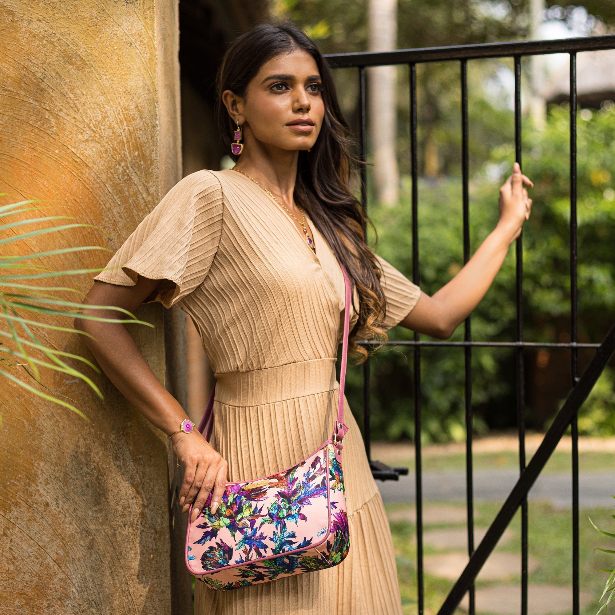 Woman standing by a gate holding an Anuschka East/West Hobo - 12013 with a main compartment, dressed in a pleated dress and accessorized with earrings and a bracelet.