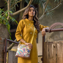 Load image into Gallery viewer, A woman in a mustard yellow outfit with double-breasted button details, holding an Anuschka Fabric with Leather Trim Crossbody with Slip Pocket - 12017 featuring an adjustable handle, poses confidently outdoors.
