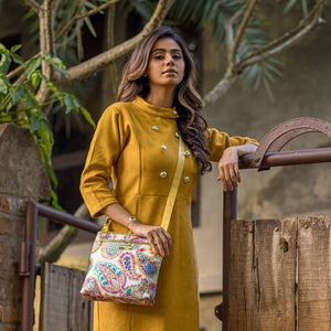 A woman in a mustard yellow outfit with double-breasted button details, holding an Anuschka Fabric with Leather Trim Crossbody with Slip Pocket - 12017 featuring an adjustable handle, poses confidently outdoors.